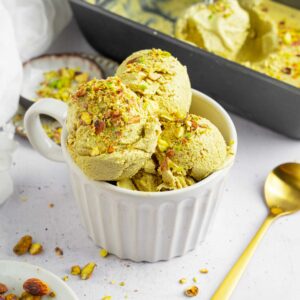 Vegan pistachio ice cream served in a white ramekin, with a golden spoon and the tub visible in the background.
