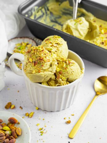 Vegan pistachio ice cream served in a white ramekin, with a golden spoon and the tub visible in the background.