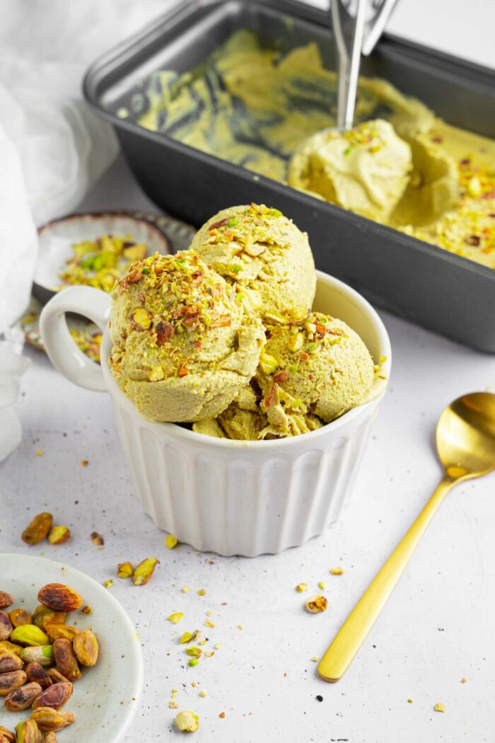 Vegan pistachio ice cream served in a white ramekin, with a golden spoon and the tub visible in the background.