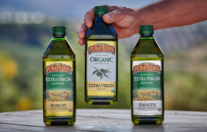 Pompeian Olive Oil products displayed with the nature backdrop, one biottle being picked up by a hand.