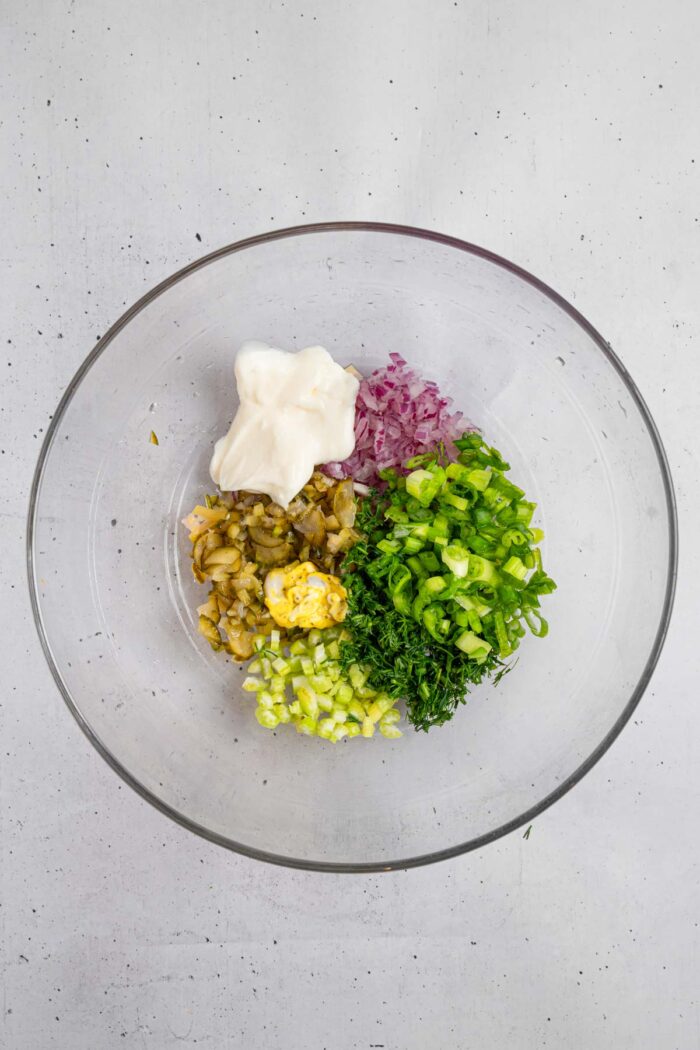 Finely chopped pickles, celery, green onions, dill and red onion, together with vegan mayo and mustard in a glass bowl.