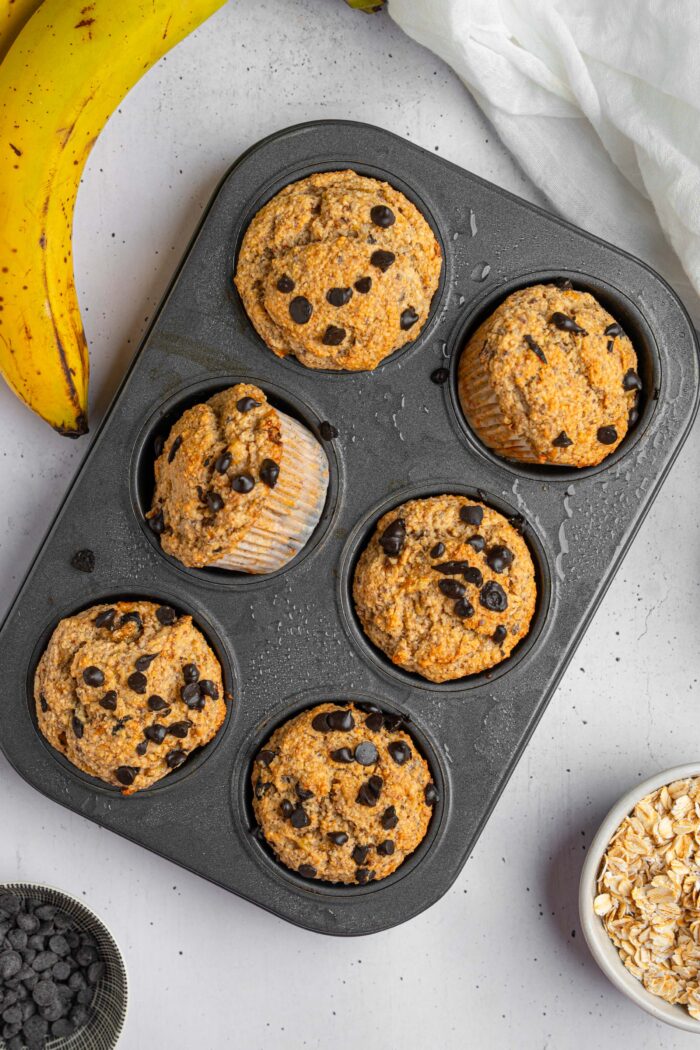 A top down view of banana oatmeal muffins in a baking tray, with ingredients such as banana and oats visible in the corners.