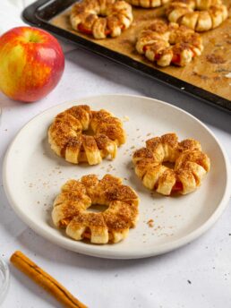 Freshly baked puff pastry apple donuts on a plate, with more visible in the background.