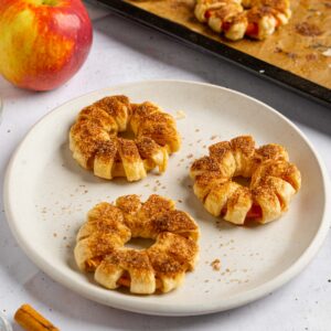 Freshly baked puff pastry apple donuts on a plate, with more visible in the background.