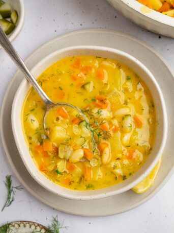 Golden creamy dill pickle soup served in a bowl, with a spoon sticking out of the soup.