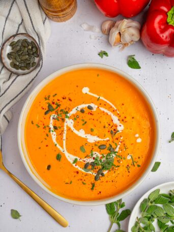 Roasted red pepper carrot soup served with a drizzle of vegan cream and some fresh herbs.