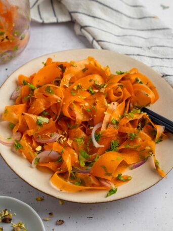 A plate full of vibrant raw carrot salad, with a jar of leftovers visible in the background.