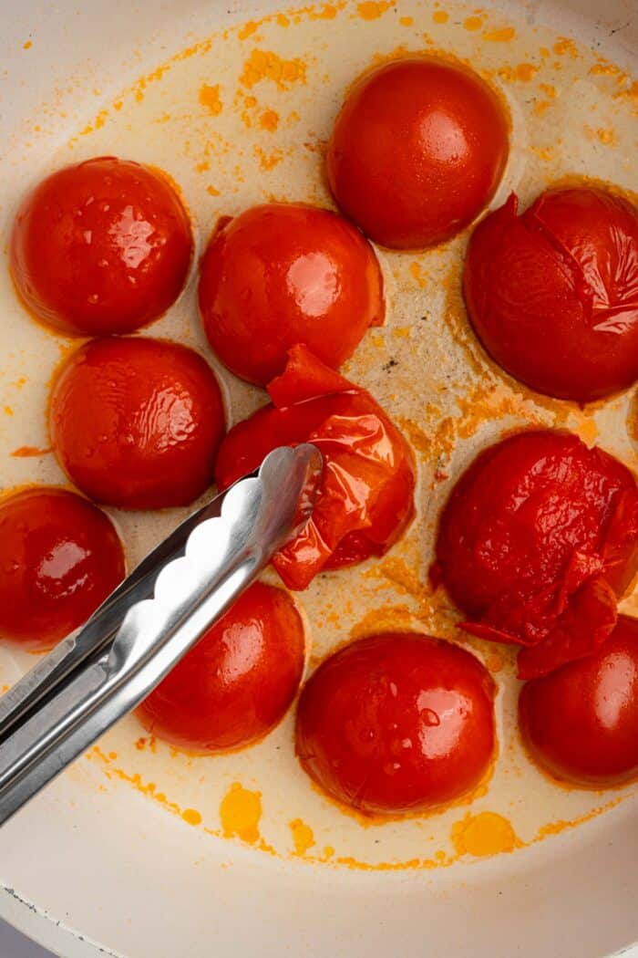 A pair of tongs pulling the skin off the Roma tomatoes while they continue cooking in the pan.