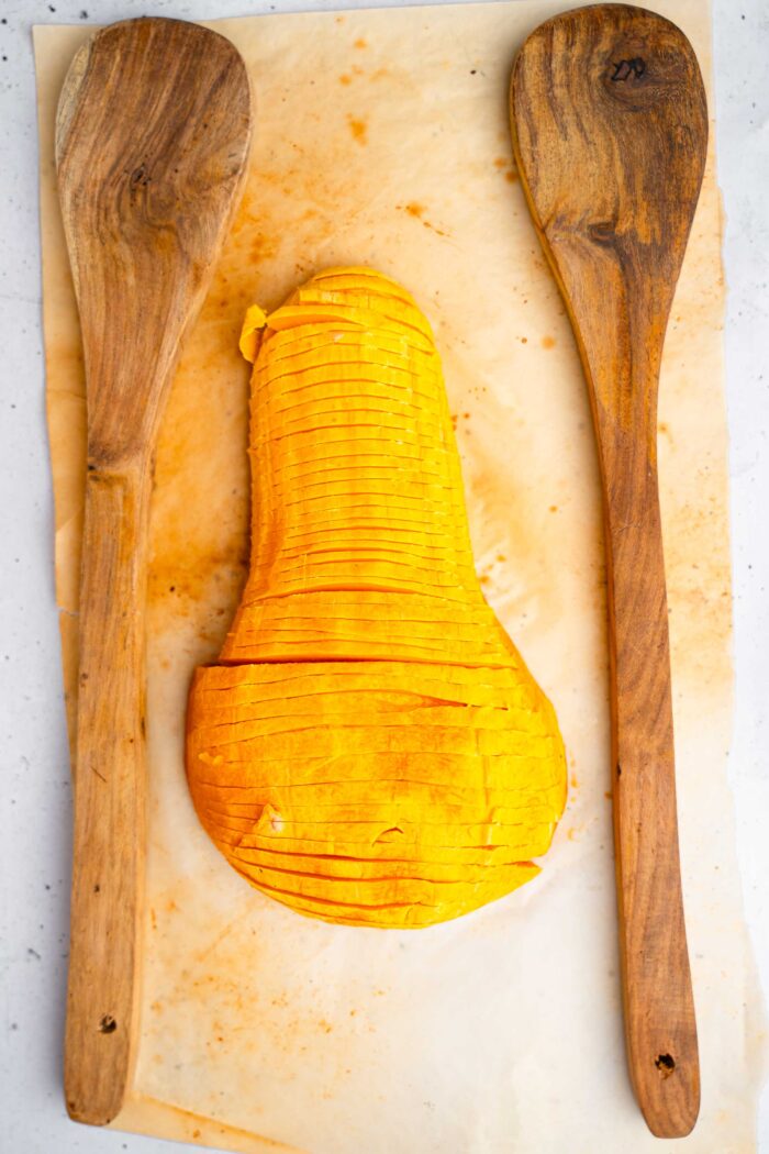 A half of roasted butternut squash cut into thin slices hasselback-style, with two wooden spoons for assistance.