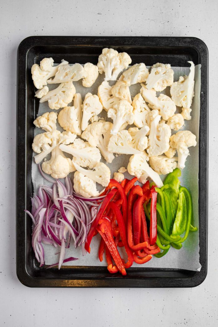 Veggies, including cauliflower, bell peppers and onions, laid out on a lined baking sheet.