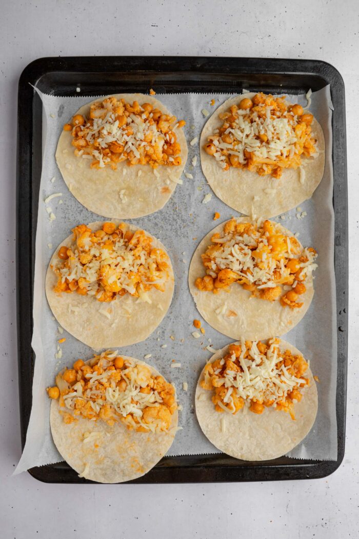 Small tortillas laid out on a lined baking sheet, with filling on half of each tortilla and some vegan cheese on top.