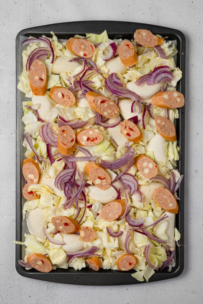The raw ingredients, including cabbage, onion, and vegan sausage, spread over the top of pierogi in a sheet pan.
