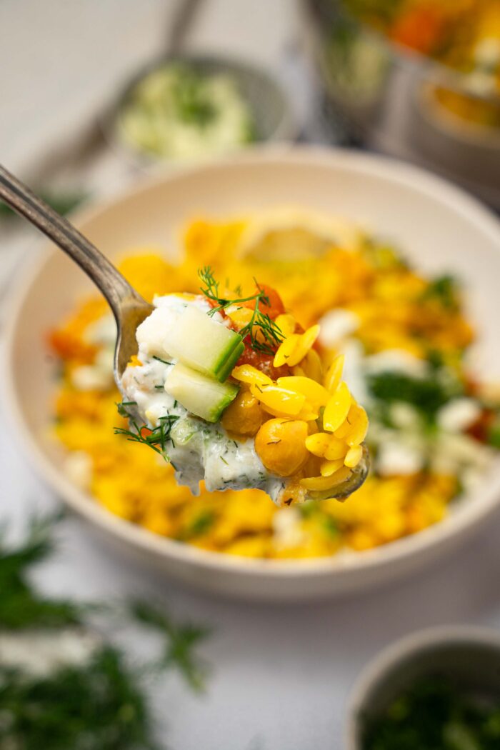 A spoonful of Mediterranean orzo bake held up to the front of the photo, with the rest ofthe bowl visible in the background.