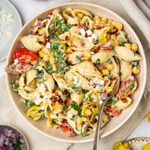 A bowl of spicy pasta salad with a spoon sticking out, and extra ingredients visible in the corners of the shot.