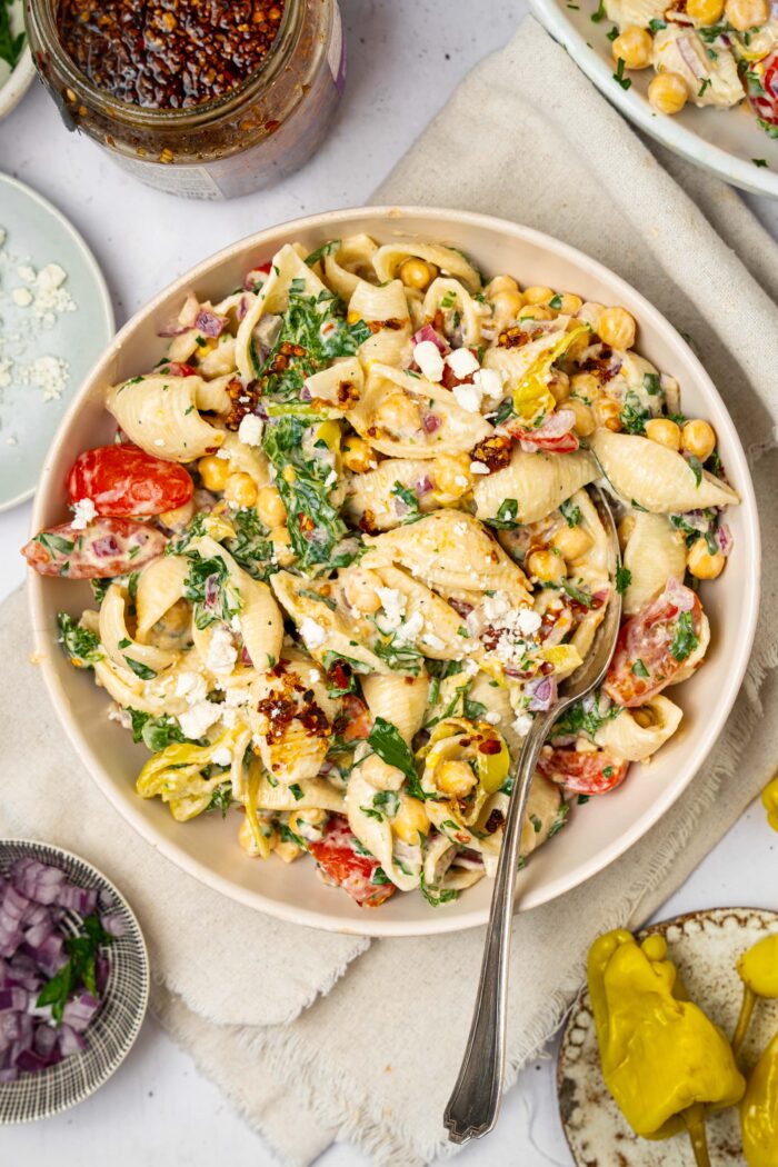 A bowl of spicy pasta salad with a spoon sticking out, and extra ingredients visible in the corners of the shot.