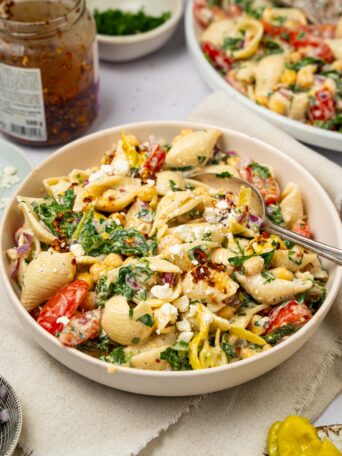 A serving of spicy pasta salad with a large salad bowl and a jar of chili crisp visible in the background.