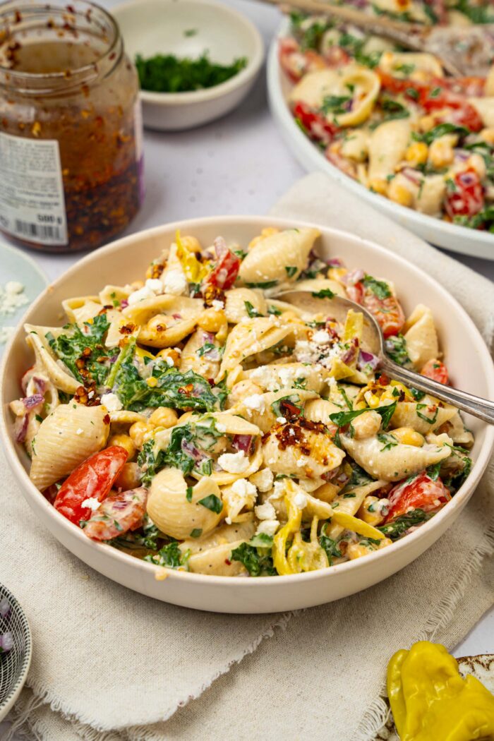 A serving of spicy pasta salad with a large salad bowl and a jar of chili crisp visible in the background.
