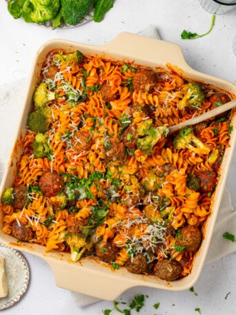 A vegan pasta and meatball bake in a square casserole dish, with a serving spoon scooping some of the food.