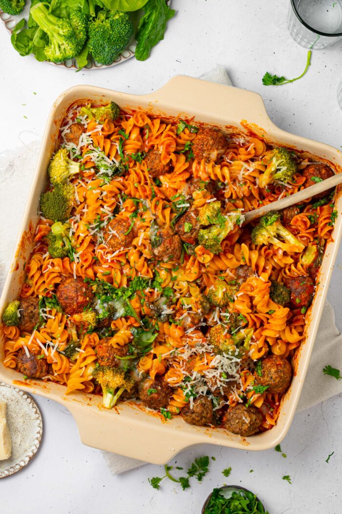 A vegan pasta and meatball bake in a square casserole dish, with a serving spoon scooping some of the food.
