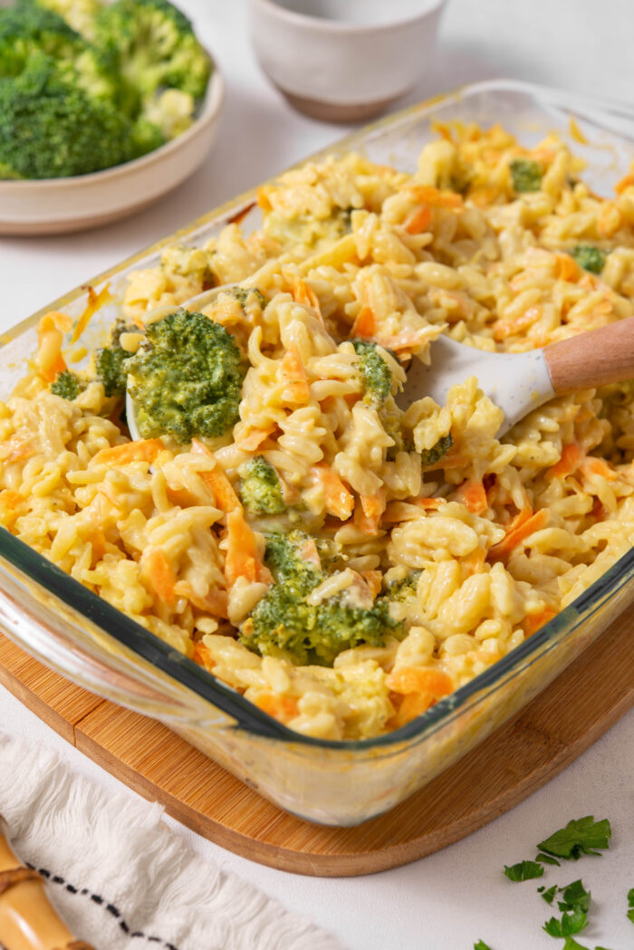 A close up of the casserole dish with vegan broccoli cheddar orzo, and a wooden serving spoon sticking out of it.