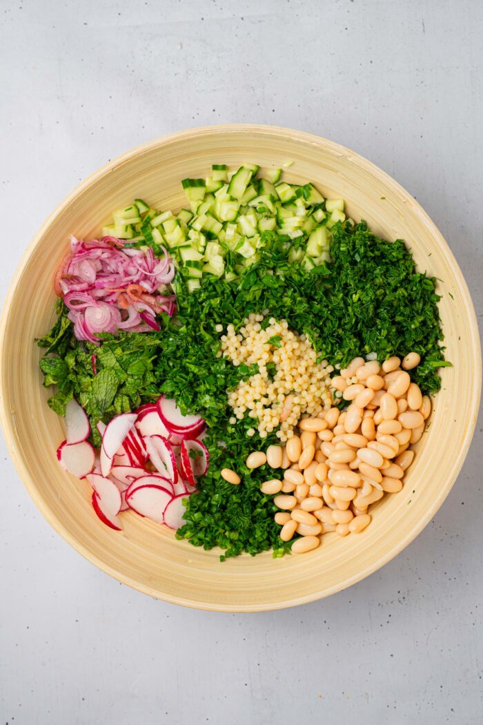 Pearl couscous, beans, greens and fresh veggies added to a large wooden mixing bowl.