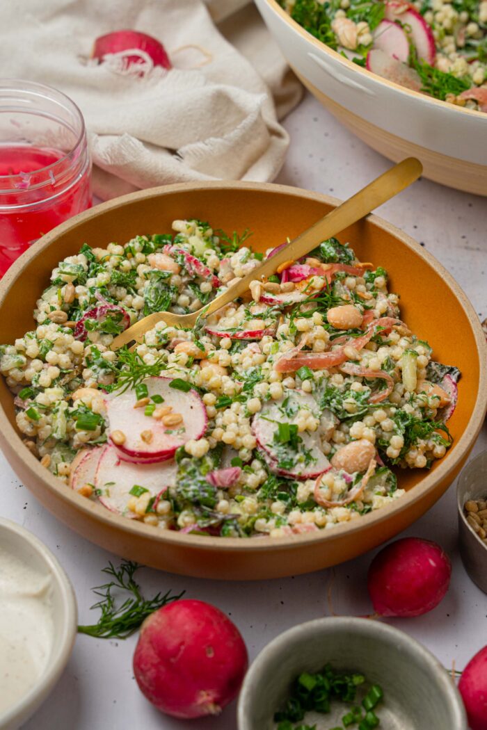 A bowl of herby ranch couscous salad served in a bowl, with different ingredients scattered around on the table.