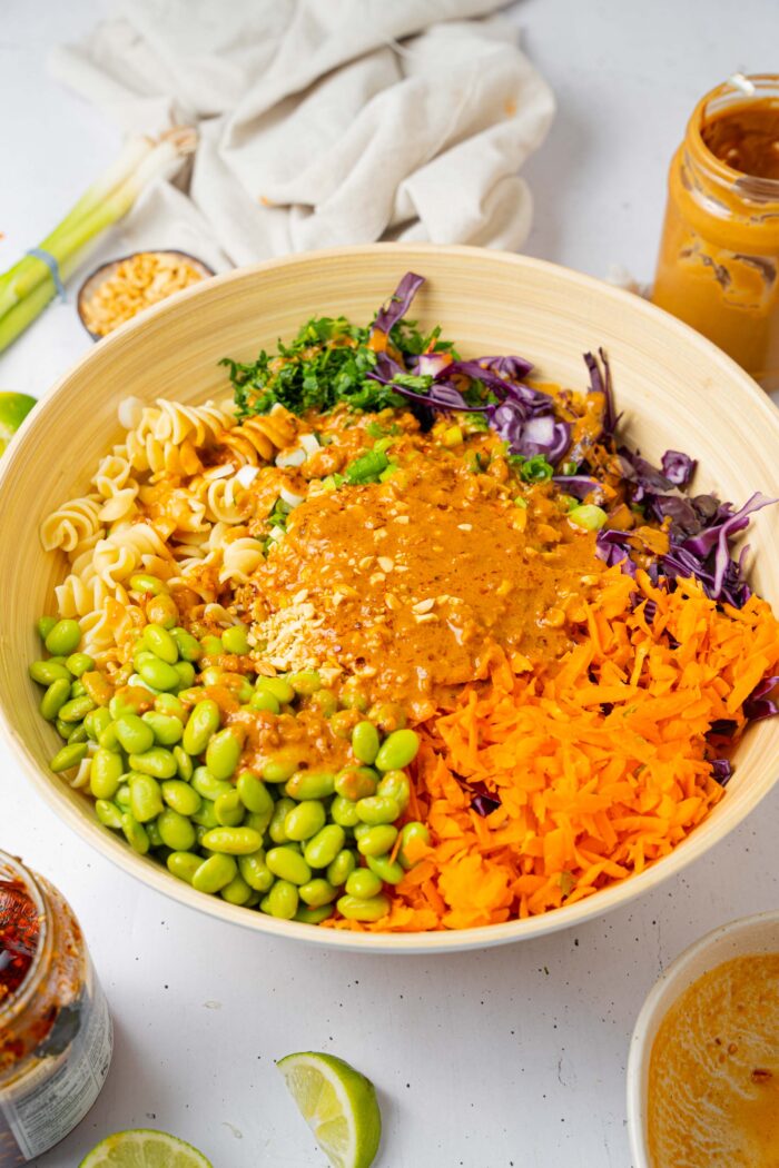 A large wooden salad bowl with various ingredients for the peanut pasta salad. including the dressing over the top.