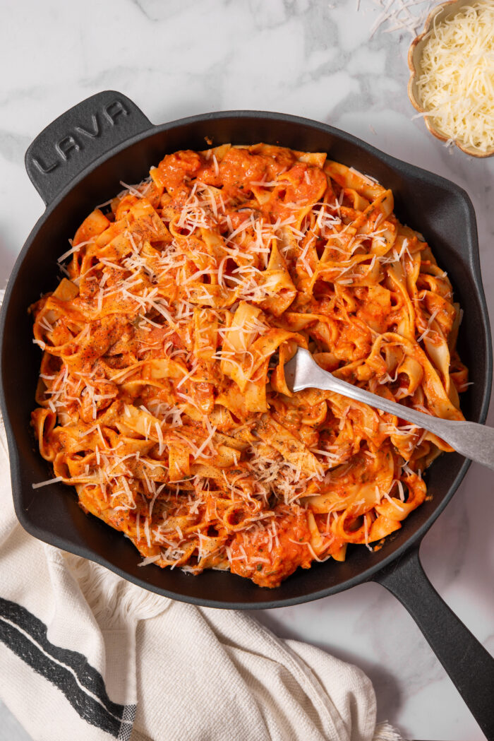 A skillet full of creamy vegan eggplant pasta, with a fork sticking out of the dish and some vegan parmesan on top.