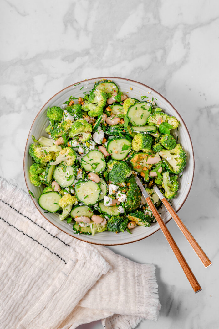 The ingredients for broccoli cucumber salad mixed together in a bowl using salad utensils.
