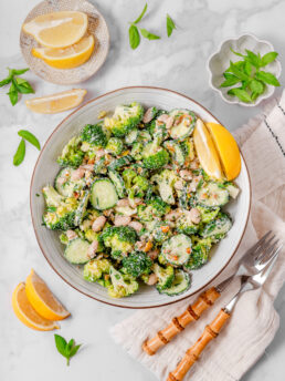 A bowl of broccoli cucumber salad with a could of forks and garnishes laid out in the peripheries.