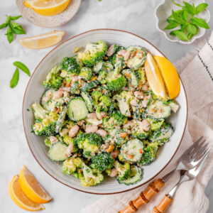 A bowl of broccoli cucumber salad with a could of forks and garnishes laid out in the peripheries.