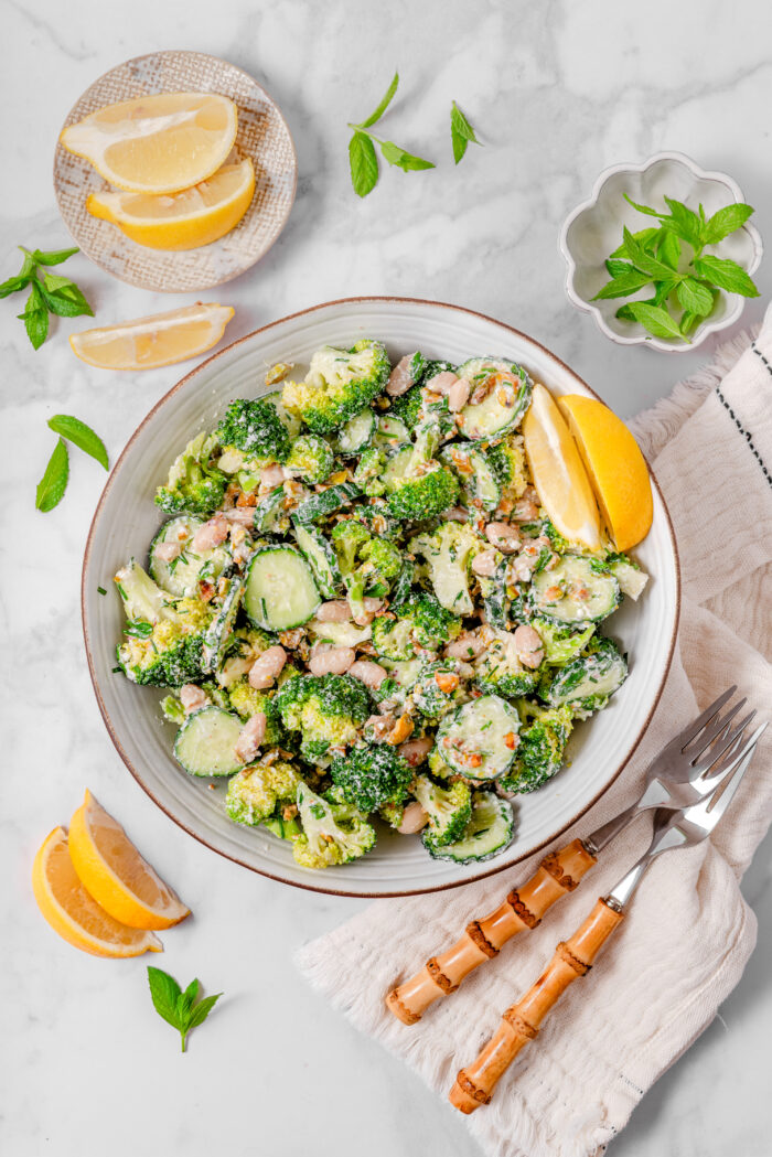 A bowl of broccoli cucumber salad with a could of forks and garnishes laid out in the peripheries.
