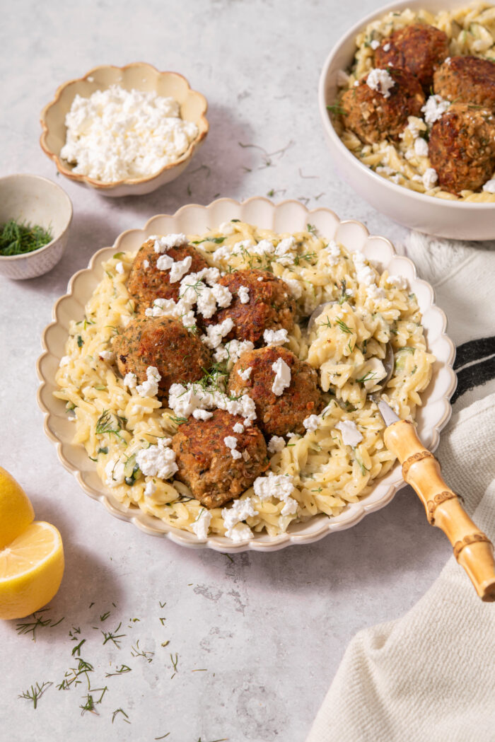 A serving of creamy zucchini orzo with vegan meatballs, fresh herbs and vegan feta.