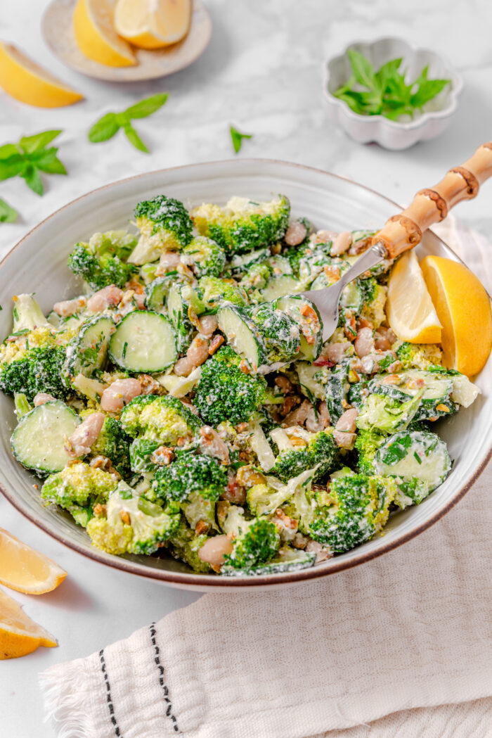 A close up of broccoli cucumber salad with a fork sticking out of the bowl, and some lemon wedges on the side.