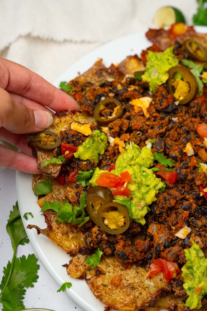 A hand picking up some loaded smashed potato nachos with a variety of toppings.