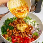 A creamy dressing with a chili swirl being poured over the crispy pasta salad.