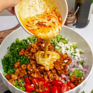A creamy dressing with a chili swirl being poured over the crispy pasta salad.