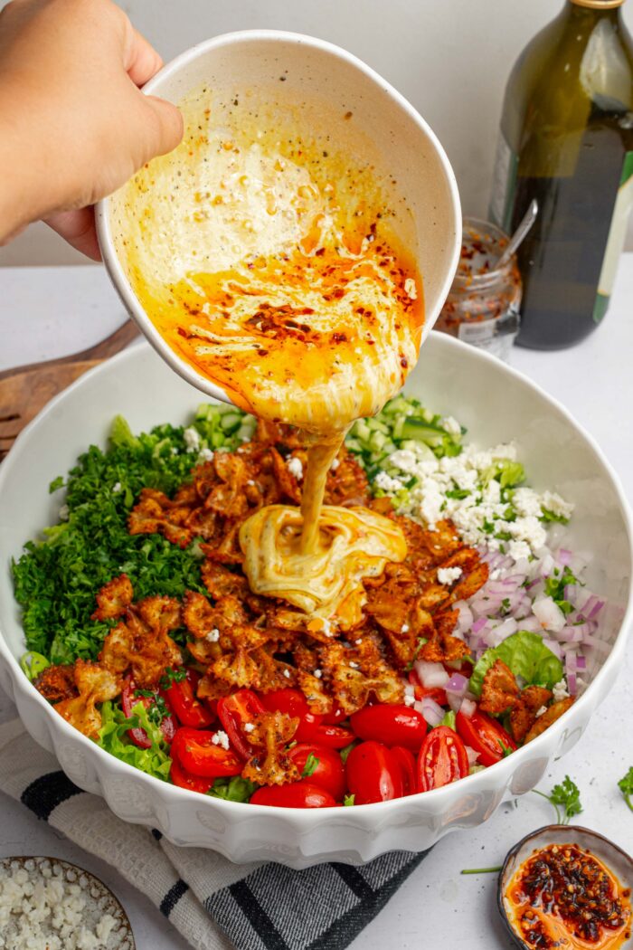 A creamy dressing with a chili swirl being poured over the crispy pasta salad.